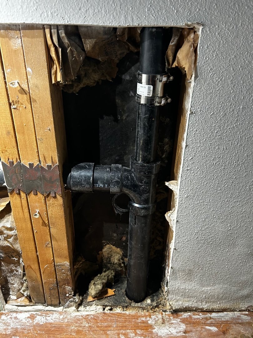 New black ABS sanitary tee and long-sweep elbow joined to the existing drain with a shielded coupling inside an opened kitchen wall