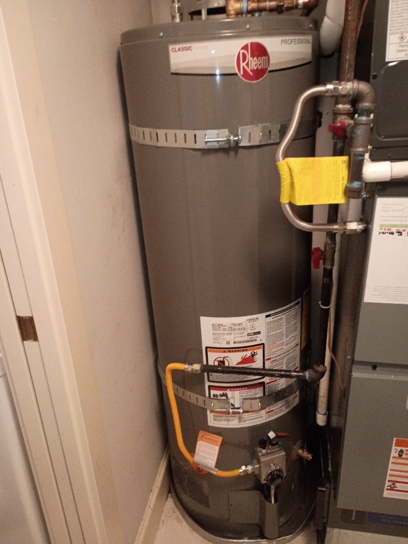 Gray tank water heater with two metal earthquake straps standing in a narrow closet beside a white door frame, with a yellow gas line at its base