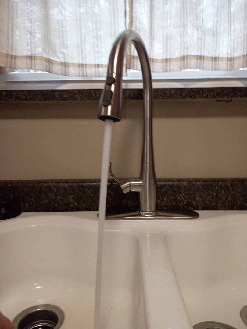 Brushed nickel pull-down kitchen faucet running water into a white double-bowl sink below a curtained window