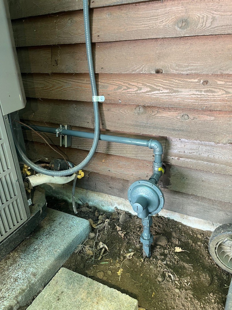 Gray steel gas pipe and a round blue-gray regulator mounted low on wood siding beside an outdoor equipment cabinet