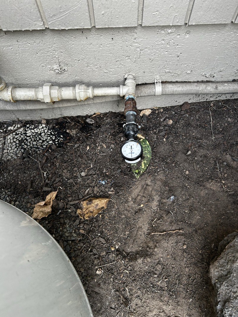 Gray steel gas pipe running along the base of light siding, with a round pressure gauge on a tee just above the soil