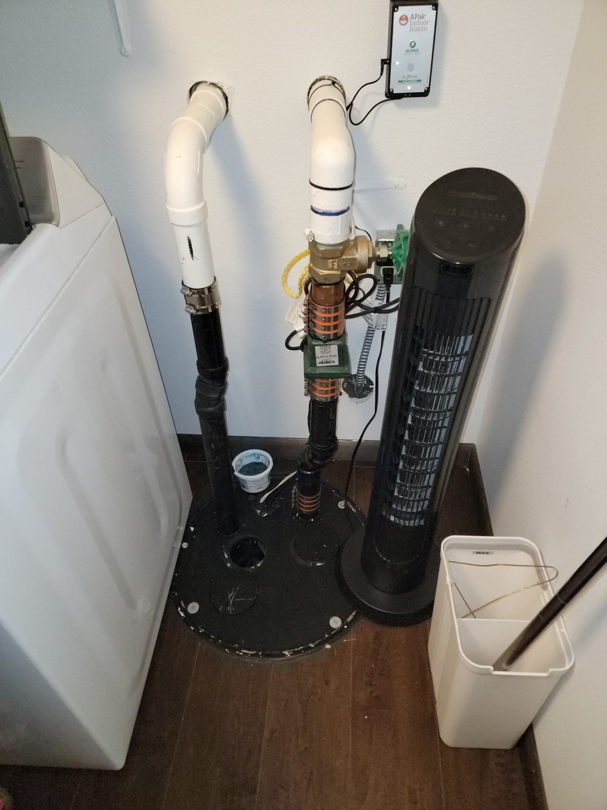 Black sealed pump basin in the corner of a laundry area, with two white PVC pipes rising from its lid and a brass valve on one of them