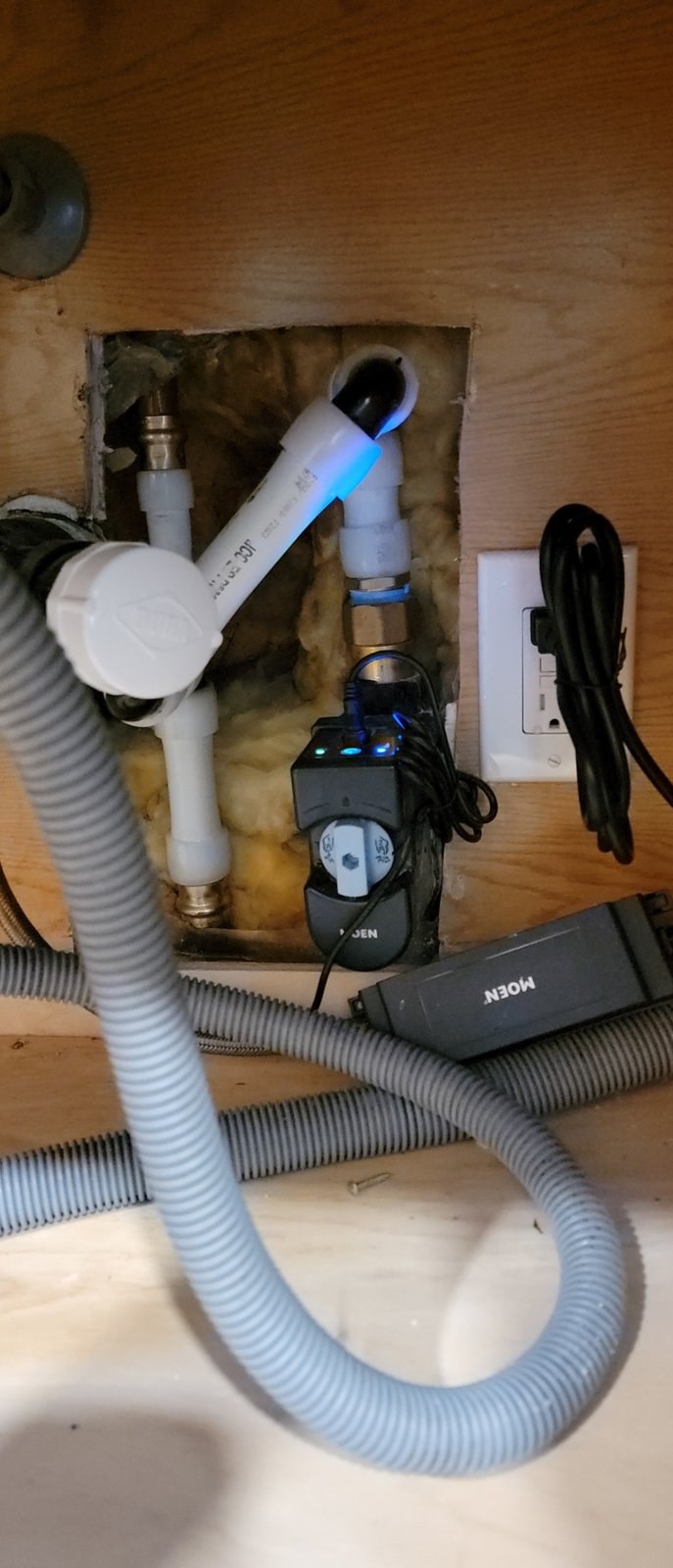 Moen Flo smart water shutoff with its indicator lights on, plumbed into white PEX with brass transition fittings inside a wall opening under a kitchen sink