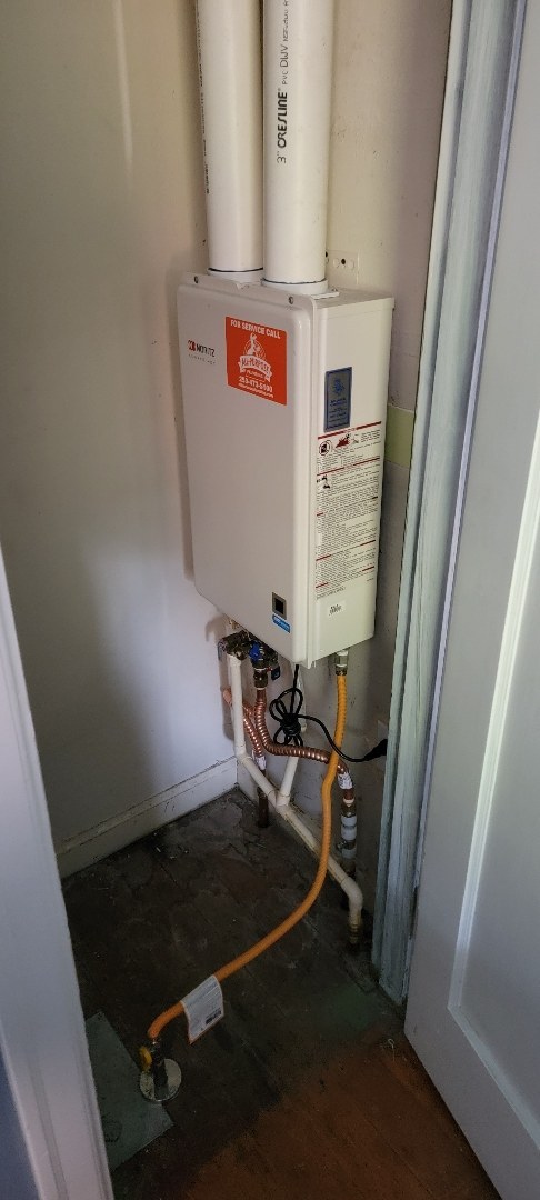 White wall-mounted tankless water heater with a red label in a narrow closet, with two white vent pipes above it and a yellow gas line below