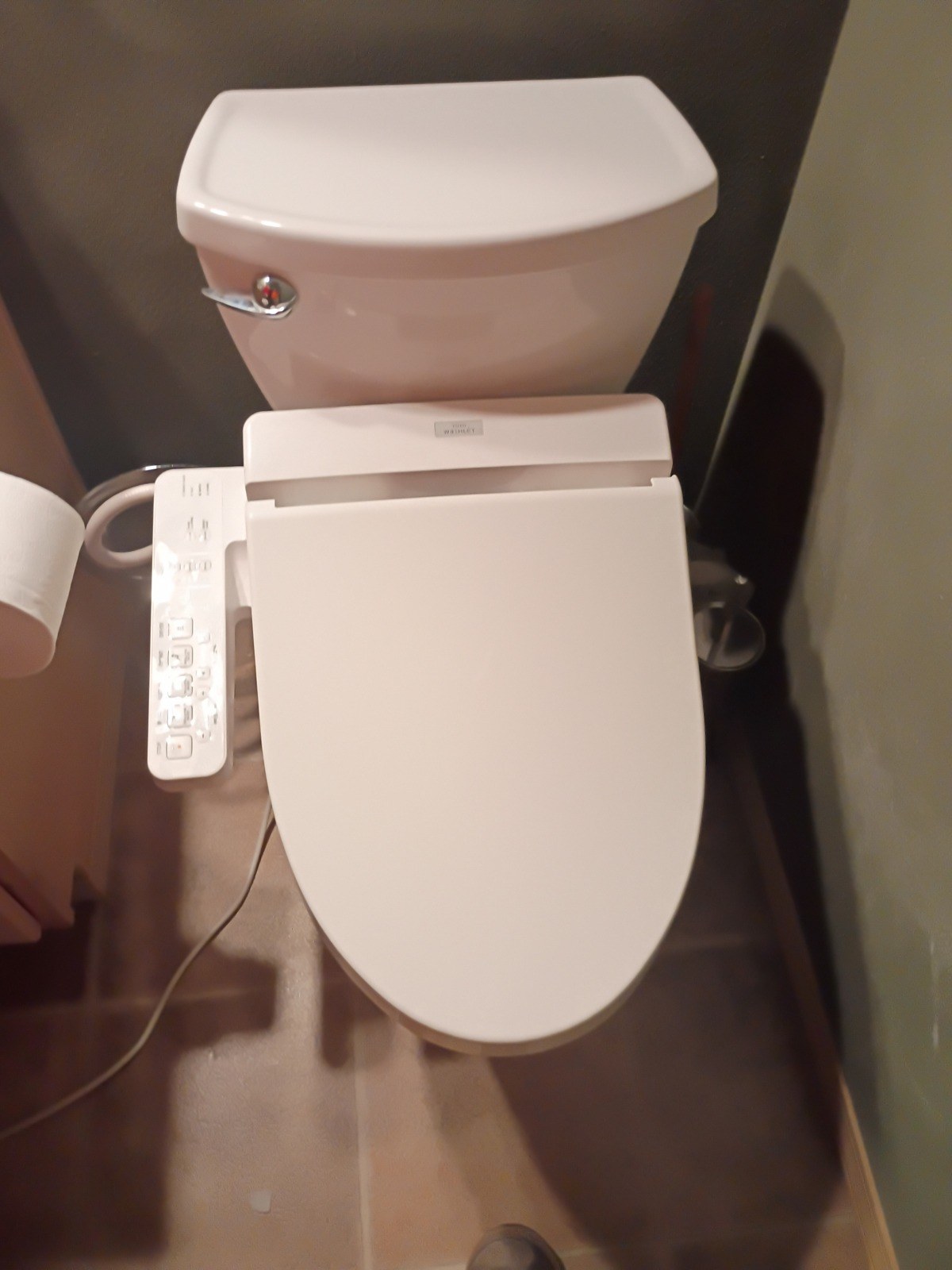 Two-piece toilet with a bidet seat and side control panel reinstalled on a tile floor against a dark wall