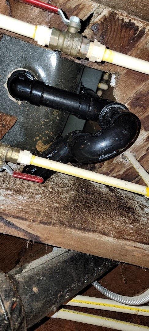 New black ABS P-trap and waste-and-overflow fitting under a bathroom floor, between the joists, with PEX supply lines and brass shutoff valves beside it