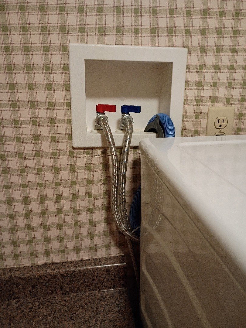 White recessed washer box with new red and blue quarter-turn valves, braided supply hoses and a blue drain hose beside a washing machine